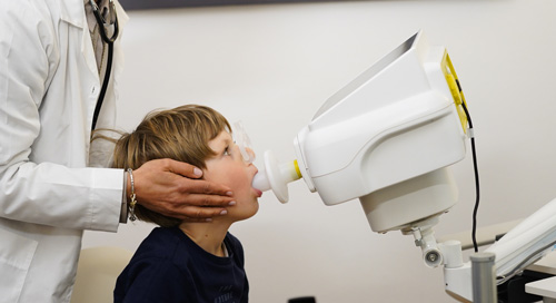 Ein Arzt (oder medizinisches Personal im weißen Kittel) hält das Mundstück des Resmon Pro V3 an den Mund eines jungen Patienten. Das Bild symbolisiert die einfache, nicht-invasive Anwendung der FOT zur Beurteilung der Atemwegsmechanik bei Kindern und älteren Patienten.